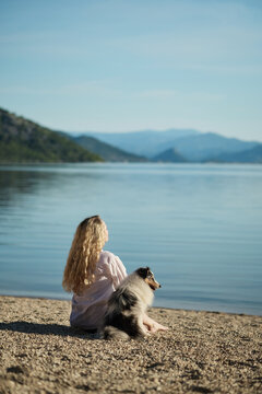 A young woman relaxes beside her Shetland Sheepdog overlooking a calm mountain lake. Peaceful outdoor lifestyle scene celebrating companionship and nature.