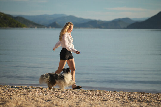 A woman enjoys a relaxing walk with her Shetland Sheepdog beside still blue water and mountains. Beautiful outdoor lifestyle image expressing freedom and wellbeing.