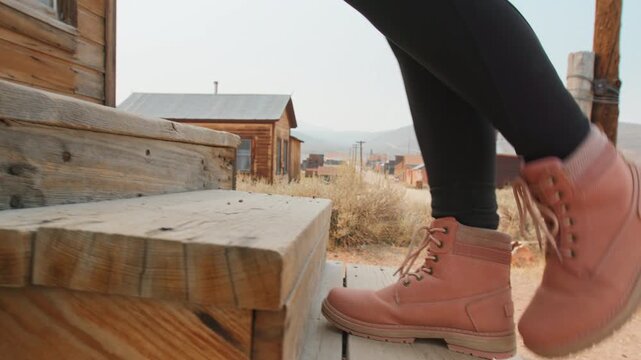 Woman wearing pink hiking boots walks along weathered wooden boardwalk in historic ghost town with rustic wooden buildings, dry desert brush, and hazy mountain backdrop under pale sky.