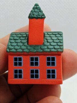 Miniature red house with green shingled roof, blue windows, and chimney held between fingers as decorative model.