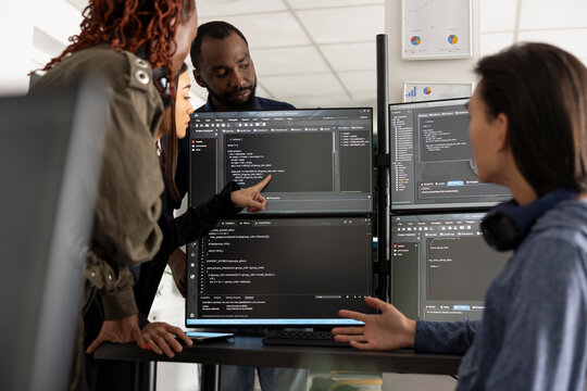 Multiethnic programmers collaborate at an office desk, reviewing code on computer screens and analyzing HTML scripts across multiple monitors.