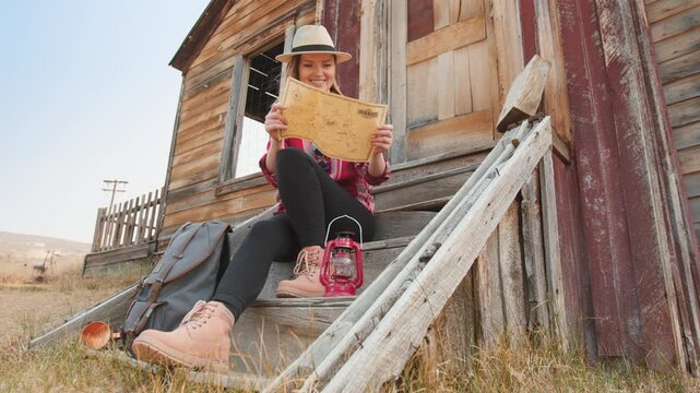 Young woman in straw hat and plaid shirt reads vintage map while sitting on weathered wooden porch of rustic abandoned building with red lantern and backpack in golden prairie grassland setting.