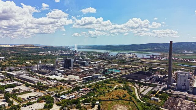 A large industrial area sits beside a river with many factories, large machinery, and roads. Clouds are visible in the blue sky as operations continue below.