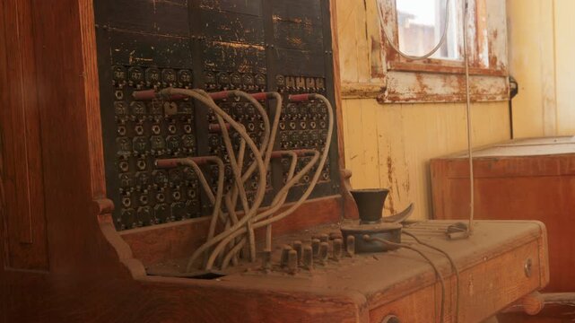 Vintage telephone switchboard with tangled patch cords and numbered connection jacks sits abandoned in dusty room with warm golden light streaming through weathered window frame and peeling paint.