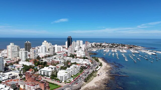 Drone view of Punta del Este Harbor, marina filled with sailboats, modern skyline and the Atlantic coastline under a clear blue summer sky in Uruguay.
