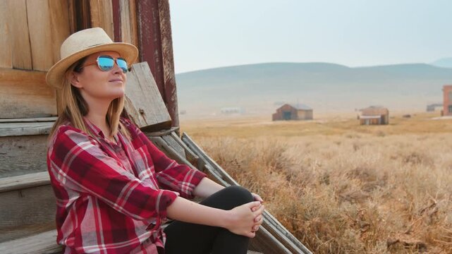 Young woman in straw hat and red plaid shirt sits peacefully on weathered wooden porch of rustic building, gazing at vast golden prairie landscape with abandoned structures under hazy mountain sky.