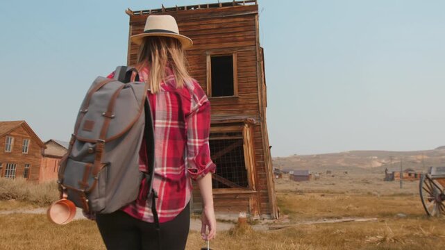 Young woman traveler wearing straw hat and plaid shirt walks toward weathered wooden buildings in historic American ghost town, golden afternoon light illuminating rustic desert landscape scenery.