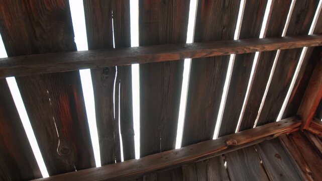 Sunlight bursts through weathered wooden slats of an old rustic barn roof, creating dramatic light rays and lens flares between aged timber planks with visible wood grain texture and rustic beams.