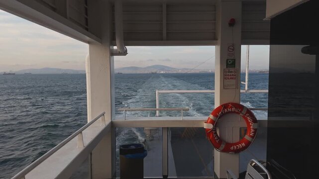 View from the deck of a moving ferry with an orange lifebuoy on a pillar and the open sea and wake visible behind during daytime, maritime travel and sea crossing scene in Turkey.