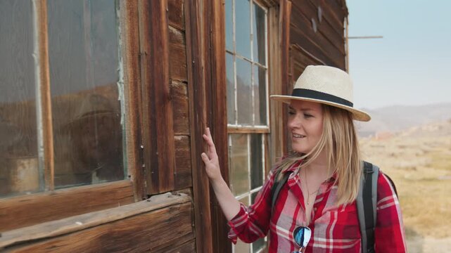 Young woman traveler wearing straw hat and red plaid shirt explores weathered wooden ghost town building.