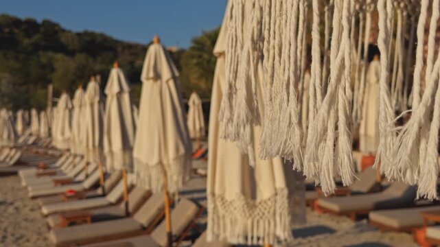 Two parallel rows of closed beige parasols and sun loungers form a symmetrical central aisle along a sunny seaside beach club, backed by green wooded hills and clear blue sky, evoking an inviting orde
