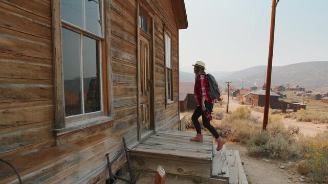 Young woman traveler in plaid shirt and straw hat explores historic ghost town, walking along weathered wooden porch of abandoned building with desert hills and rustic structures in warm hazy light.
