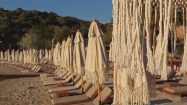 A long diagonal row of closed beige parasols and sun loungers recedes into the distance along a sunny seaside resort shore, framed by green wooded hills and clear blue sky, creating a striking perspec