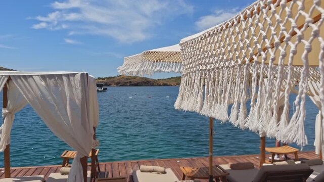 Close-up shot of a fringed macrame umbrella and white cabana drapes on a wooden beach deck, overlooking a turquoise bay and distant green hills.
