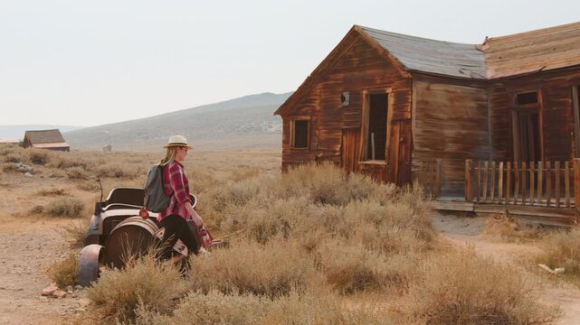 Young woman traveler wearing plaid shirt and straw hat explores historic abandoned ghost town with weathered wooden cabins, rusty vintage car remains, dry desert sagebrush.