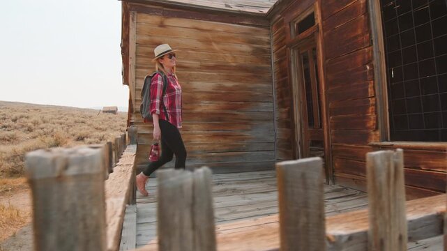 Girl walks through old abandoned ghost town in America exploring weathered wooden buildings. Female traveler in plaid shirt and straw hat discovers historic western architecture with desert landscape.