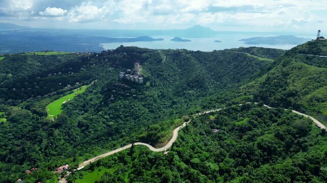 Wide establishing drone shot of Tagaytay Highlands featuring Woodridge Place, People's Park in the Sky road, tropical forests, Taal Lake, and Taal Volcano during bright daytime.