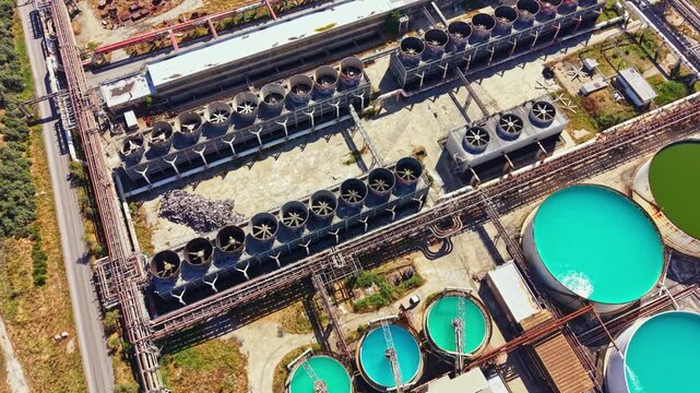 An industrial site shows large circular tanks filled with bright green liquid. Several cooling towers stand nearby. The area has some gravel paths and machinery around it. It is daytime.