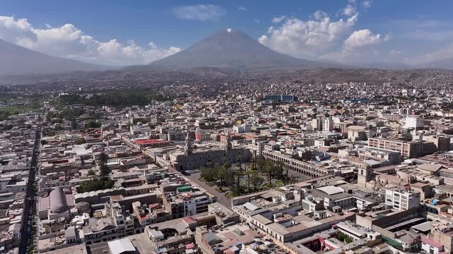 Aerial drone footage of Arequipa, Peru, featuring the iconic Misti volcano towering over Plaza de Armas and the historic city center on a clear sunny day. Scenic urban landscape in the Peruvian Andes.
