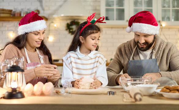 Christmas family baking cookies in kitchen. Parents and daughter knead dough at home, wearing Santa hats and antlers amid warm holiday lights. Celebration of family holiday traditions.