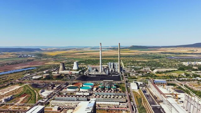 A large industrial site features tall smokestacks and cooling towers. Nearby, there are water pools and open land. The scene includes a river and agricultural fields in the background.