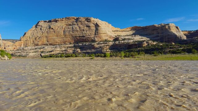 Harding Hole backcountry camping overlook spot, rippled sand foreground with towering canyon cliffs and desert vegetation beyond, harsh morning light, pan right