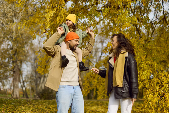 Happy family together in autumn park with daughter. Parents walk under golden leaves while child rides on dad's shoulders, and mom gently steadies them. Family bonding outdoors in happiness.