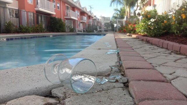 A broken glass sits by a pool, fragments scattered alongside the brick pathway