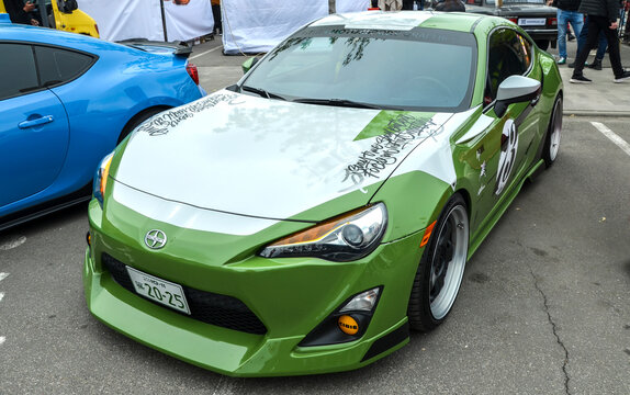 Green Scion FR-S sports coupe in an olive livery with white and grey vinyl graphics. Lowered suspension, custom deep-dish wheels, and modified aero kit at a car meet