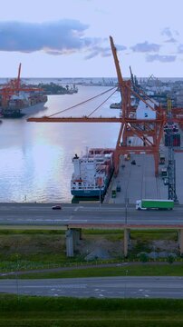 Cinematic aerial shot of the Gdynia mobile port in Poland featuring container ships, industrial cranes, and traffic on the estakada bridge during golden hour.