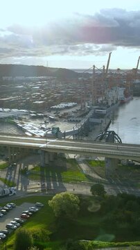 A cinematic aerial shot showing the industrial harbor in Gdynia, Poland, featuring cargo cranes, shipping containers, and traffic on the Estakada bridge during sunset.