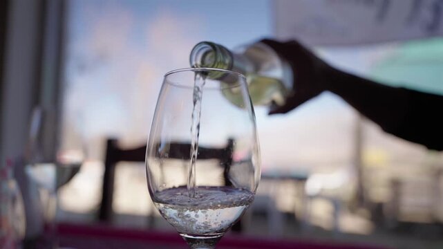 White wine poured from a glass bottle into a stemmed wine glass on a restaurant table with a softly blurred dining setting in the background, close-up, static shot.