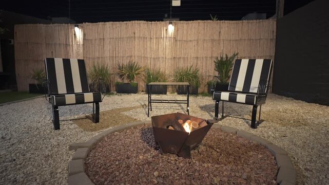 A relaxing night view of a modern backyard patio. A warm fire burns in a metal fire pit surrounded by gravel, black and white striped chairs, and an illuminated bamboo fence.
