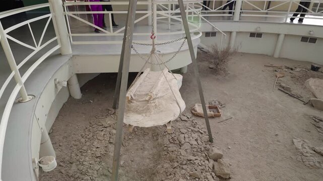 A large dinosaur fossil encased in a protective plaster jacket hangs from a metal tripod hoist at an excavation site in Ischigualasto Provincial Park, Argentina.