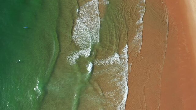 Waves flow onto the sandy beach under a clear sky in the coastal area during daylight hours aerial view. Shoreline shows patterns made by water on the sand.
