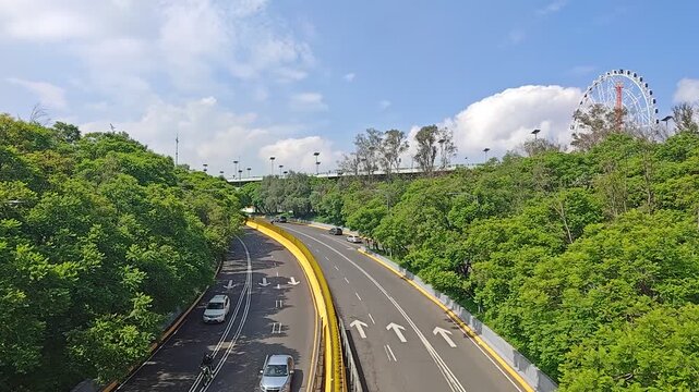 Static shot of winding road through Bosque de Chapultepec with cars passing and Ferris wheel in background, Mexico City