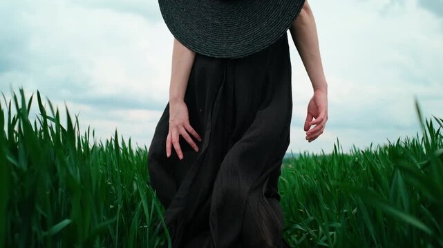 Low rear view of woman walking away through saturated green field, wearing matte black dress and round wide brim hat; flowing fabric, visible back slit, soft overcast light, calm outdoor fashion mood