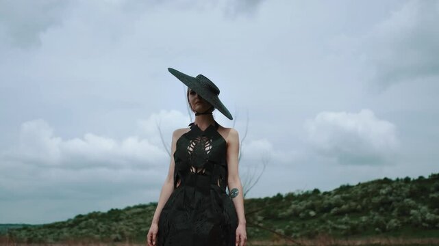 Centered low-angle portrait of solitary woman in open countryside under heavy overcast sky, wearing wide-brim hat and textured cutout dress, creating moody commercial fashion mood, with woman, model