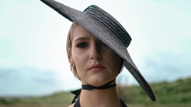 Close coastal portrait shows young woman in wide brim woven hat tilting face slightly left, brim throwing diagonal shadow across one eye while ribbon choker rests beneath chin in soft natural light