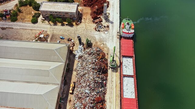 A ship is loading scrap metal at a dock next to a storage facility. Piles of scrap are visible on the ground. The scene takes place during daylight hours.