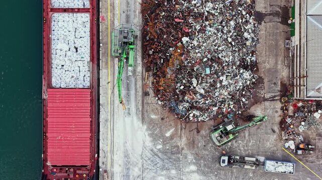 A cargo ship unloads materials at a recycling center. Heavy machines work to move and sort large piles of scrap metal and debris. Workers are present to manage the operations.