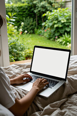 Naklejka premium Woman types on modern laptop with blank white screen sitting on bed. Freelancer works at home in bedroom. View of backyard garden with green plants through glass door. Person uses portable electronic