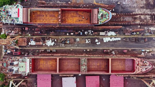 Workers are engaged in shipbuilding at a shipyard. Two large vessels are visible, with sections of the ships showing the structure being assembled. Tools and materials are scattered around.
