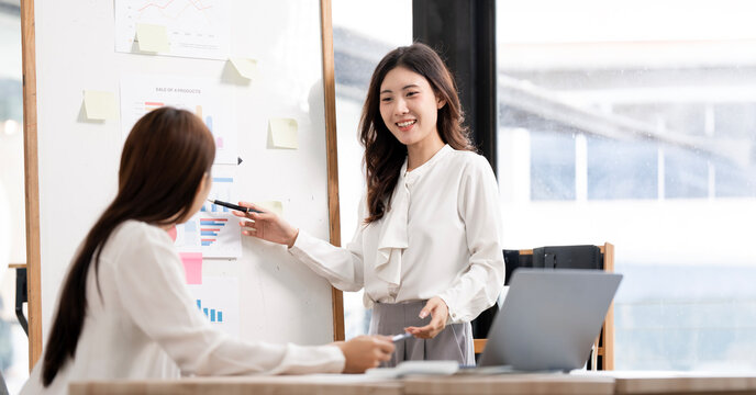Young Asian businesswomen discussing financial charts on whiteboard in office.
