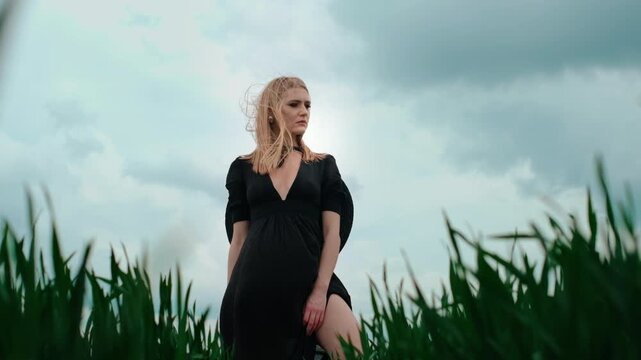 Blonde woman wearing black dress with deep V-neck and ribbon choker stands in tall green grass viewed from low-angle viewpoint; hair and skirt gently lifted by breeze, hand resting on exposed knee