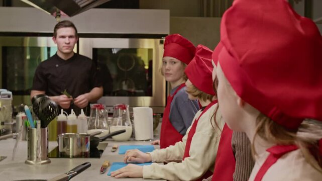 Caucasian teenagers wearing red chef hats listening to male chef instructor during culinary class. Footage is ideal for promoting children cooking workshops and educational programs