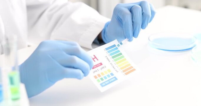 Scientist holding PH test strip with color table at white table in laboratory, closeup
