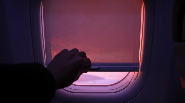 Hand opening airplane window shade to reveal colorful sunset sky