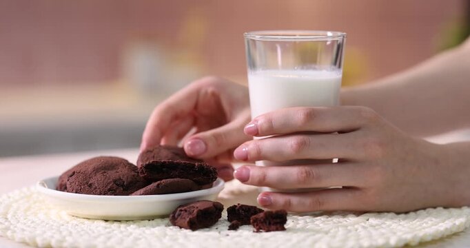 Woman putting glass of fresh milk onto table and taking tasty chocolate cookie from plate in kitchen, closeup