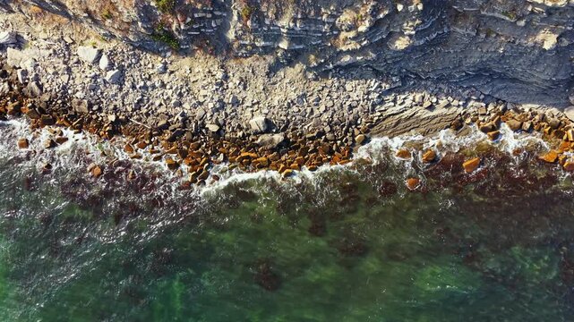 Waves hit the rocky shore while water remains clear. Stones and pebbles are visible under the sunlight. The coastline stretches out in the background as a natural scene unfolds.